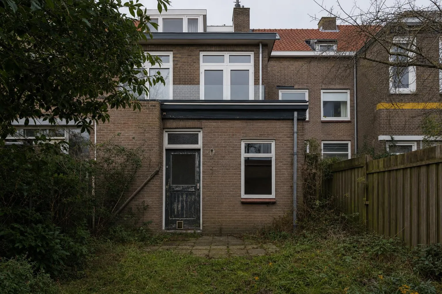 Dezelfde woning vóór de uitbouw: gedateerde achtergevel met verweerde deur en verwilderde tuin