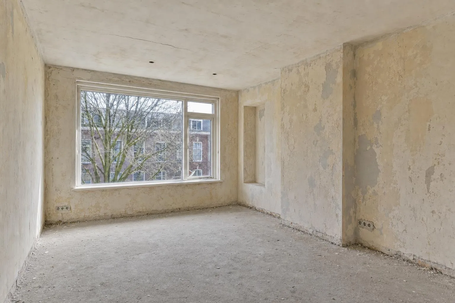 Dezelfde kamer vóór het stucwerk: gescheurde, vlekkerige muren en een kale betonvloer